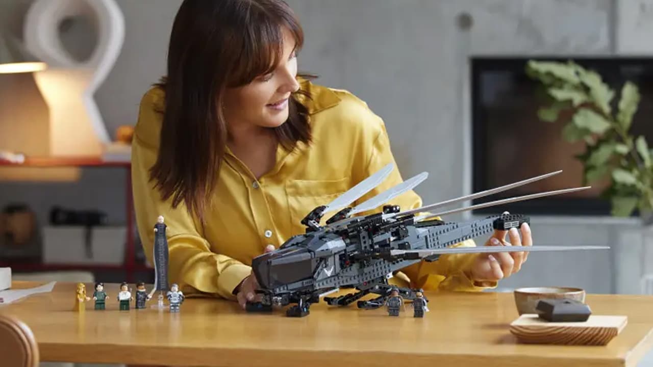 A woman in a yellow shirt holding a Dune Lego Ornithopter set, with the minifigures on the desk.